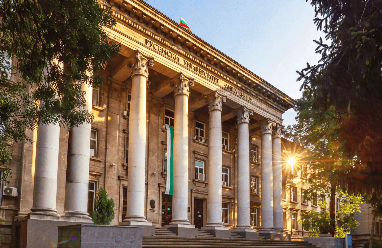 A historic building with ancient columns is illuminated by the evening sun. The Bulgarian flag flies on the roof as well as above the main entrance. There are stairs and trees in front of the building.