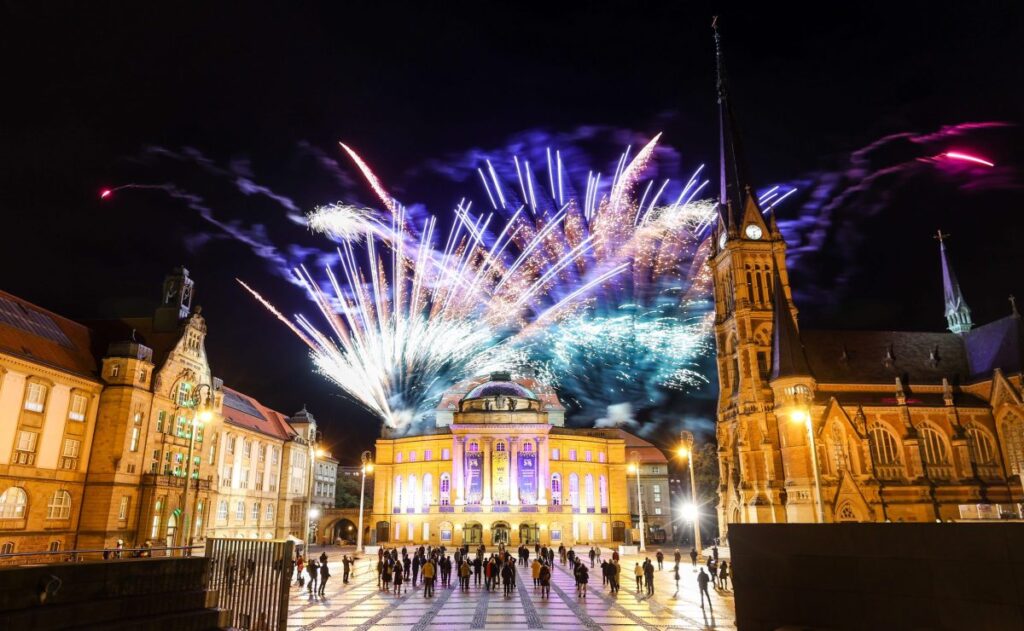 Chemnitz Theatre Square. A three-part ensemble of buildings on a large square. In the centre is a neo-renaissance opera house behind which a colourful firework display can be seen. The firework as a symbol of joint forces of the European Universities contributing to the European Universities Week.