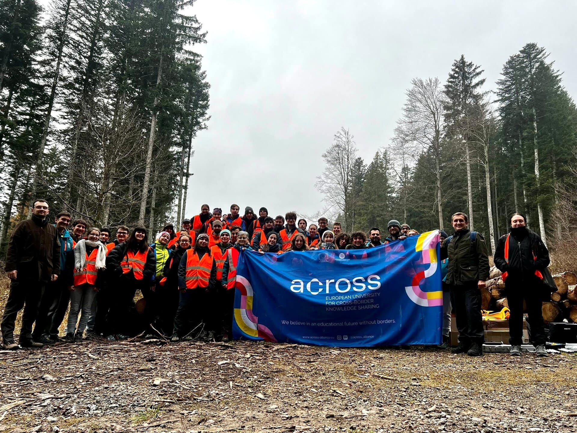 Around fifty students from the University of Udine and University of Banja Luka posing in Pramosio Forest with a large Across Alliance banner after completing an environmental monitoring weekend.